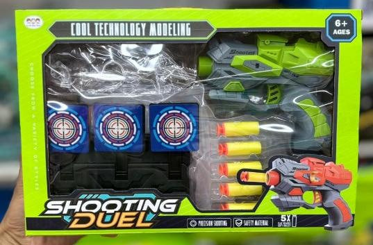 A boxed toy shooting game set labeled 'Cool Technology Modeling' and 'Shooting Duel' in bright neon green packaging, containing a green and gray toy gun, three blue target discs, and four yellow foam darts with orange tips. A hand is holding the package.