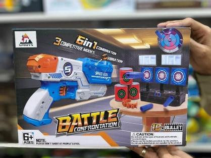 A person holds a boxed toy blaster set labeled 'Battle Confrontation' in a retail store. The packaging shows a blue, white, and orange toy gun, target boards, and small red and blue accessories.