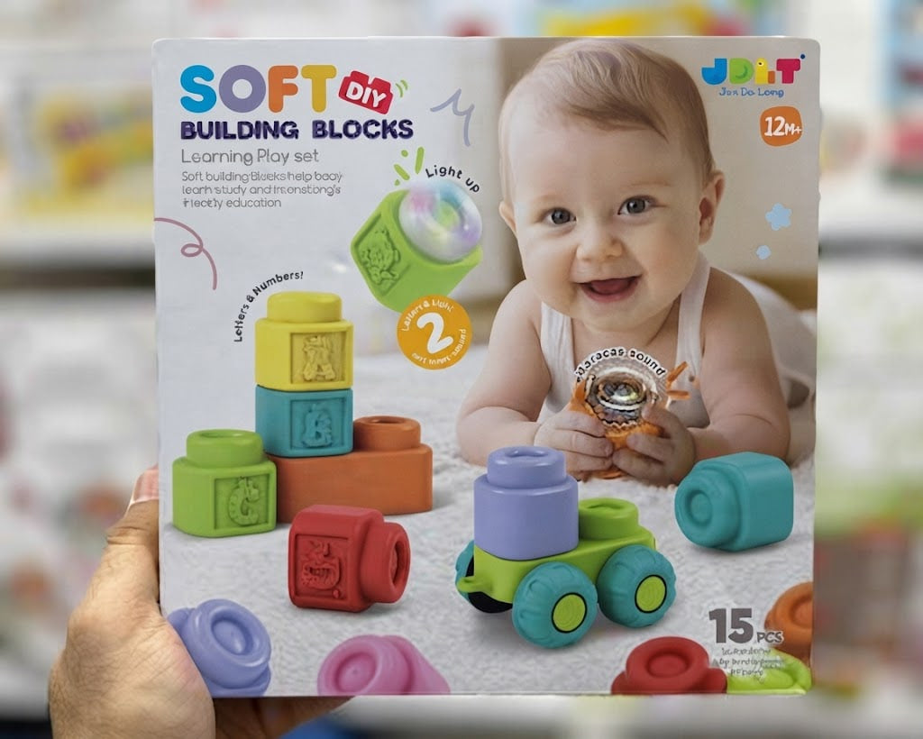 A hand holds a boxed toy set labeled 'SOFT DIY BUILDING BLOCKS' with a smiling baby pictured on the package. The box shows colorful building blocks and a small train toy, with the brand name JOY+T visible in the top right corner.