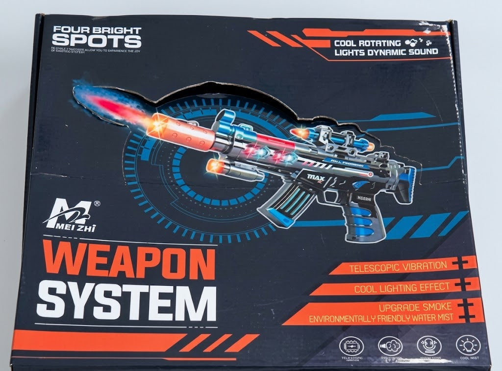 Product packaging box for a toy weapon system, featuring a black background with blue and orange graphics, a printed toy gun illustration, and text including 'FOUR BRIGHT SPOTS' and 'WEAPON SYSTEM'.