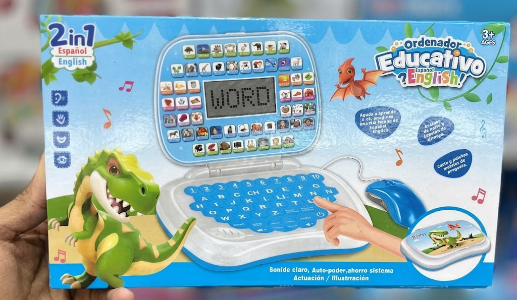 A hand holding a 2-in-1 educational English toy laptop package. The box features a blue and white laptop design with a dinosaur illustration, a mouse, and Portuguese text including 'Ordenador Educativo English'.