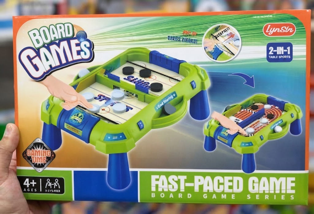 A hand holds a LynSin board games box featuring a green and blue 2-in-1 tabletop game set, with printed graphics of the game table and sports icons on the packaging.