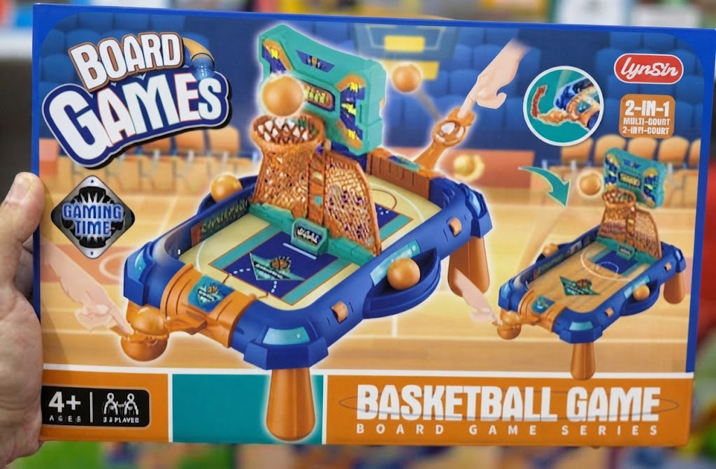 A hand holds a LynSir board game box featuring a colorful basketball game set. The packaging shows a blue and orange table game with basketball hoops, balls, and basketball court graphics, with the text 'BASKETBALL GAME BOARD GAME SERIES' and age rating '4+'.