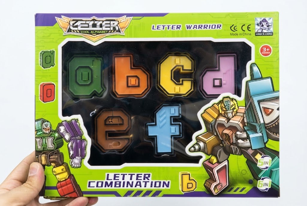 A hand holding a boxed toy set labeled 'Letter Warrior' and 'Letter Combination' with colorful alphabet-shaped pieces visible through the front window. The packaging is bright green with cartoon robot illustrations and includes age and certification marks.